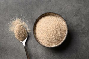 a wooden spoon with a pile of psyllium husk powder next to a bowl filled with Plantago ovata soluble fiber