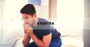 a man sitting on a bed holding his head, appearing distressed, possibly experiencing symptoms like fatigue or emotional strain.