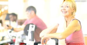 a smiling woman in her 40s using an exercise bike in a gym, demonstrating active lifestyle and cvičení for hubnutí.