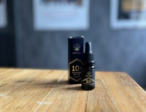 a 10% full-spectrum CBD oil dropper bottle from CBDmylife with 10 ml, 1000 mg CBD, placed beside its black packaging on a wooden surface.